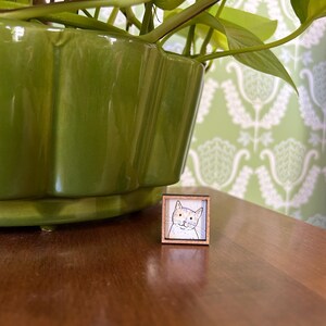 May include: A small, square wooden cat portrait with a light brown frame. The cat is white with orange and black markings, and a black outline. The artwork is on a wooden surface, with a green plant pot and patterned wallpaper in the background.