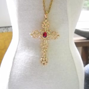May include: A gold-toned metal cross pendant suspended from a matching gold-toned chain. The cross features an intricate, openwork filigree design with a single, oval-shaped, deep red gemstone set prominently in the center of the cross piece.