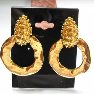 May include: Gold-toned earrings with a textured, clustered top and a hammered, open-circle design. The earrings are displayed on a black card, showcasing their bright, reflective surface. These earrings are a statement piece.