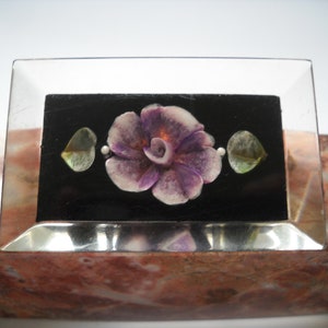 May include: A purple and white flower with green leaves is set against a black background inside a clear glass frame.