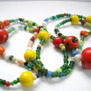Op de afbeelding: Een meerstrengige ketting met groene, witte, rode, gele en blauwe kralen. De ketting heeft grote, ronde, rode kralen met witte en zwarte beschilderde ontwerpen.
