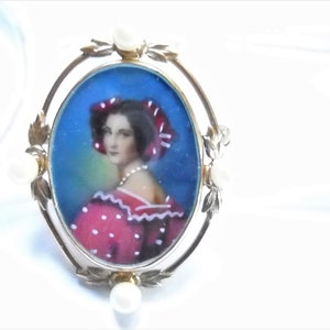 May include: A vintage gold-toned brooch with a painted portrait of a woman in a red and white polka dot dress. The portrait is set in an oval frame with four pearl accents.