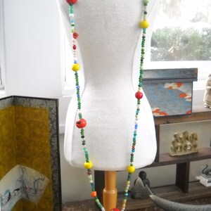 Op de afbeelding: Een kleurrijke kralenketting met rode, gele, groene en blauwe kralen. De ketting is op een wit manikin en wordt gepresenteerd op een houten standaard.