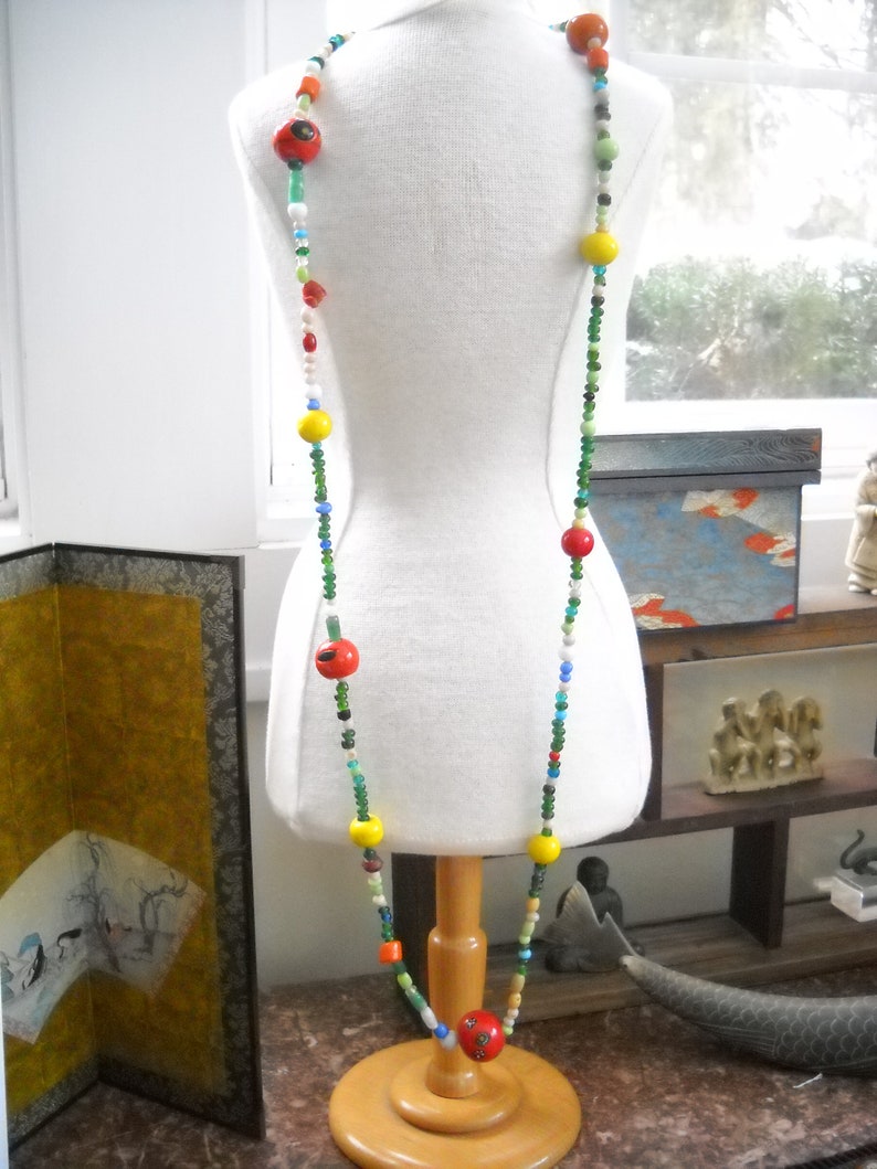 Op de afbeelding: Een kleurrijke kralenketting met rode, gele, groene en blauwe kralen. De ketting is op een wit manikin en wordt gepresenteerd op een houten standaard.