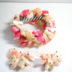 K&ouml;nnte beinhalten: Ein Set aus rosa und wei&szlig;en Muschel-Schmuck, bestehend aus einem Armband und einem Paar Ohrringen. Das Armband besteht aus einem Metallband, an dem eine Reihe von rosa und wei&szlig;en Muschelteilen befestigt sind. Die Ohrringe bestehen ebenfalls aus rosa und wei&szlig;en Muschelteilen.