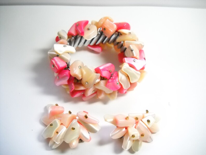 K&ouml;nnte beinhalten: Ein Set aus rosa und wei&szlig;en Muschel-Schmuck, bestehend aus einem Armband und einem Paar Ohrringen. Das Armband besteht aus einem Metallband, an dem eine Reihe von rosa und wei&szlig;en Muschelteilen befestigt sind. Die Ohrringe bestehen ebenfalls aus rosa und wei&szlig;en Muschelteilen.