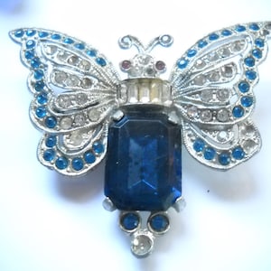 May include: A silver-toned butterfly brooch featuring a large rectangular dark blue central stone, accented with smaller round blue and clear rhinestones along the wings and body, and two small red stones near the head of the insect design.