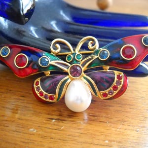 May include: A decorative butterfly brooch with red, blue, green, and purple enamel wings. The gold-coloured metal brooch features a large faux pearl and is decorated with various coloured rhinestones.