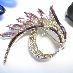 May include: A silver tone brooch with a swirl design. The brooch is decorated with clear and purple rhinestones.