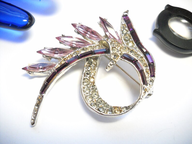 May include: A silver tone brooch with a swirl design. The brooch is decorated with clear and purple rhinestones.