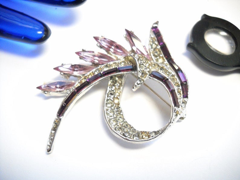 May include: A silver tone brooch with a swirling design featuring clear and pink rhinestones. The brooch has a pin back.