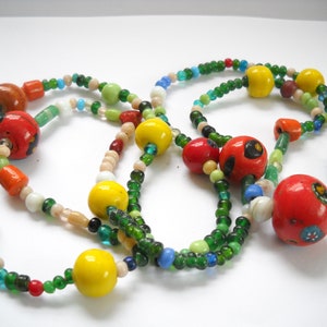 Op de afbeelding: Een meerstrengige ketting met groene, gele, rode, oranje, witte en blauwe kralen. De ketting heeft grotere, ronde, rode kralen met beschilderde ontwerpen.