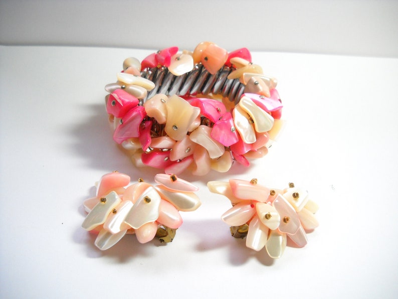 K&ouml;nnte beinhalten: Ein Set aus rosa und wei&szlig;en Muschel-Schmuck, bestehend aus einem Armband und einem Paar Ohrringen. Das Armband besteht aus einem Metallband mit einer Ansammlung von rosa und wei&szlig;en Muschelteilen. Die Ohrringe sind ebenfalls aus rosa und wei&szlig;en Muschelteilen gefertigt und haben einen Clipverschluss.