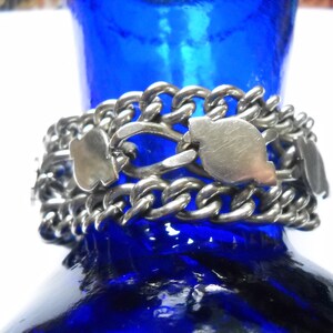 May include: A silver chain bracelet with multiple links and decorative charms. The bracelet is made of a thick, chunky chain and features several silver charms in the shape of hearts. The bracelet is resting on a blue glass surface.