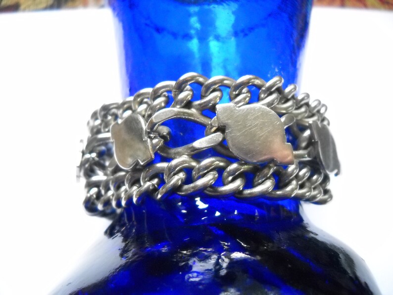 May include: A silver chain bracelet with multiple links and decorative charms. The bracelet is made of a thick, chunky chain and features several silver charms in the shape of hearts. The bracelet is resting on a blue glass surface.