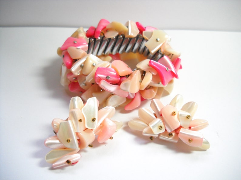 K&ouml;nnte beinhalten: Ein Armband und Ohrringe Set aus rosa und wei&szlig;en Muscheln. Das Armband besteht aus einem Metallband, an dem rosa und wei&szlig;e Muschelteile befestigt sind. Die Ohrringe bestehen ebenfalls aus rosa und wei&szlig;en Muschelteilen.