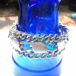May include: A silver chain bracelet with heart-shaped charms. The bracelet is displayed on a blue glass bottle.
