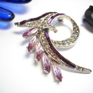 May include: A silver tone brooch with a swirl design. The brooch is decorated with clear and pink rhinestones and purple baguette-shaped stones.