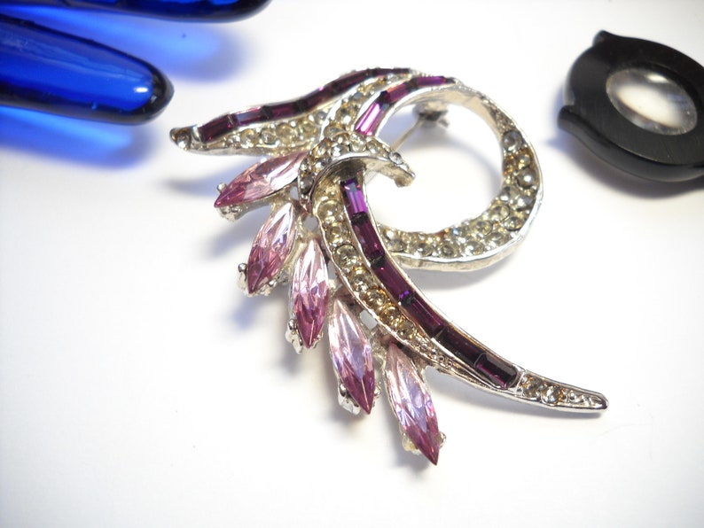 May include: A silver tone brooch with a swirl design. The brooch is decorated with clear and pink rhinestones and purple baguette-shaped stones.