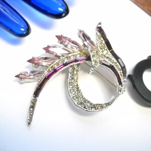 May include: A silver tone brooch with a swirl design featuring pink and purple rhinestones.
