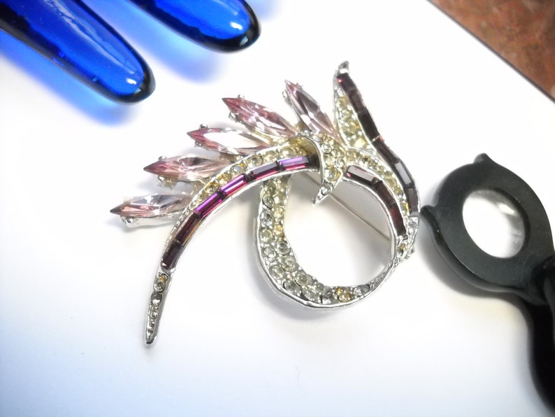 May include: A silver tone brooch with a swirl design featuring pink and purple rhinestones.