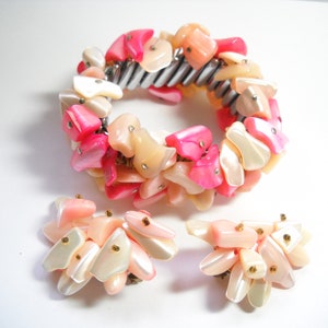 K&ouml;nnte beinhalten: Ein rosa-wei&szlig;es Muschel-Armband und passende Ohrringe. Das Armband besteht aus einem Metallband, an dem eine Reihe von rosa-wei&szlig;en Muschelteilen befestigt sind. Die Ohrringe bestehen ebenfalls aus rosa-wei&szlig;en Muschelteilen.