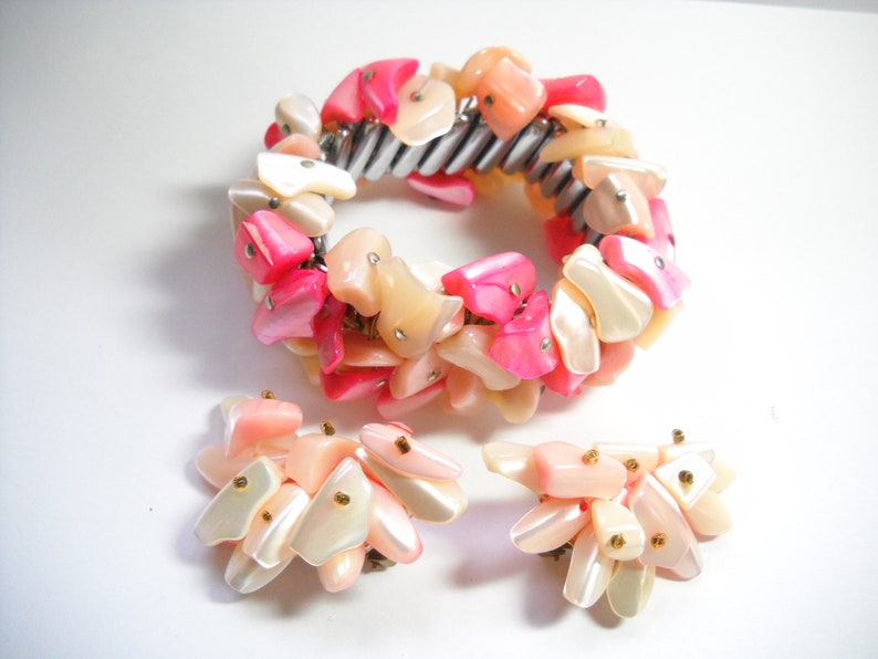 K&ouml;nnte beinhalten: Ein rosa-wei&szlig;es Muschel-Armband und passende Ohrringe. Das Armband besteht aus einem Metallband, an dem eine Reihe von rosa-wei&szlig;en Muschelteilen befestigt sind. Die Ohrringe bestehen ebenfalls aus rosa-wei&szlig;en Muschelteilen.
