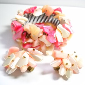 K&ouml;nnte beinhalten: Ein pinkfarbenes und wei&szlig;es Muschel-Armband und passende Ohrringe. Das Armband besteht aus einer Reihe kleiner, unregelm&auml;&szlig;ig geformter Muscheln, die auf einem Metallband aufgereiht sind. Die Ohrringe bestehen ebenfalls aus kleinen, unregelm&auml;&szlig;ig geformten Muscheln, die an einem Metallstift befestigt sind.