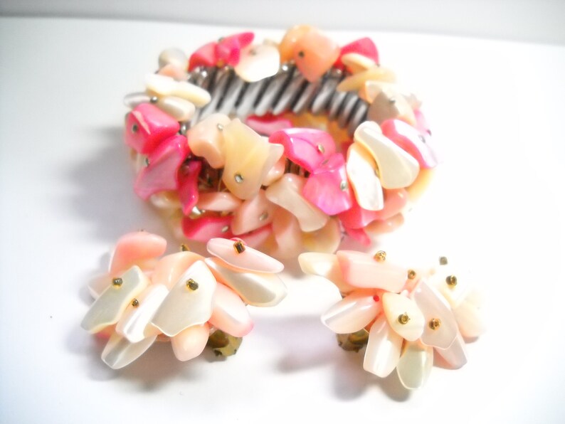 K&ouml;nnte beinhalten: Ein pinkfarbenes und wei&szlig;es Muschel-Armband und passende Ohrringe. Das Armband besteht aus einer Reihe kleiner, unregelm&auml;&szlig;ig geformter Muscheln, die auf einem Metallband aufgereiht sind. Die Ohrringe bestehen ebenfalls aus kleinen, unregelm&auml;&szlig;ig geformten Muscheln, die an einem Metallstift befestigt sind.