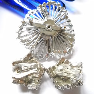 May include: A silver-toned brooch and a pair of clip-on earrings with clear rhinestones. The brooch is shaped like a sunburst and the earrings are square.