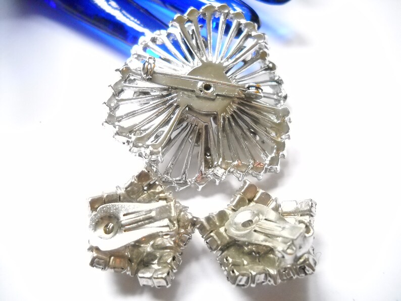 May include: A silver-toned brooch and a pair of clip-on earrings with clear rhinestones. The brooch is shaped like a sunburst and the earrings are square.
