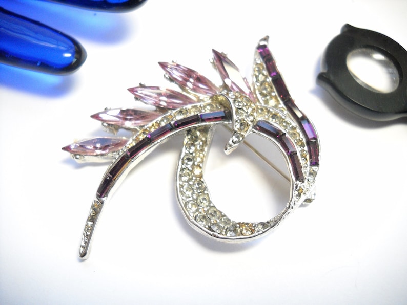 May include: A silver tone brooch with a swirling design featuring clear and pink rhinestones. The brooch has a long, thin, pointed shape with a cluster of pink rhinestones at the top.