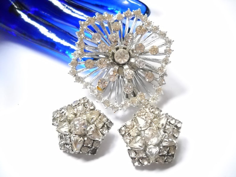May include: A set of vintage rhinestone jewelry featuring a large brooch with a starburst design and two matching earrings. The jewelry is silver-toned and features clear rhinestones.