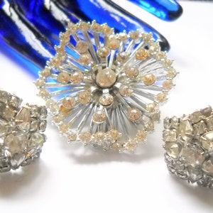 May include: A silver tone brooch and matching earrings set with clear rhinestones. The brooch is a starburst design with a central rhinestone. The earrings are star shaped.