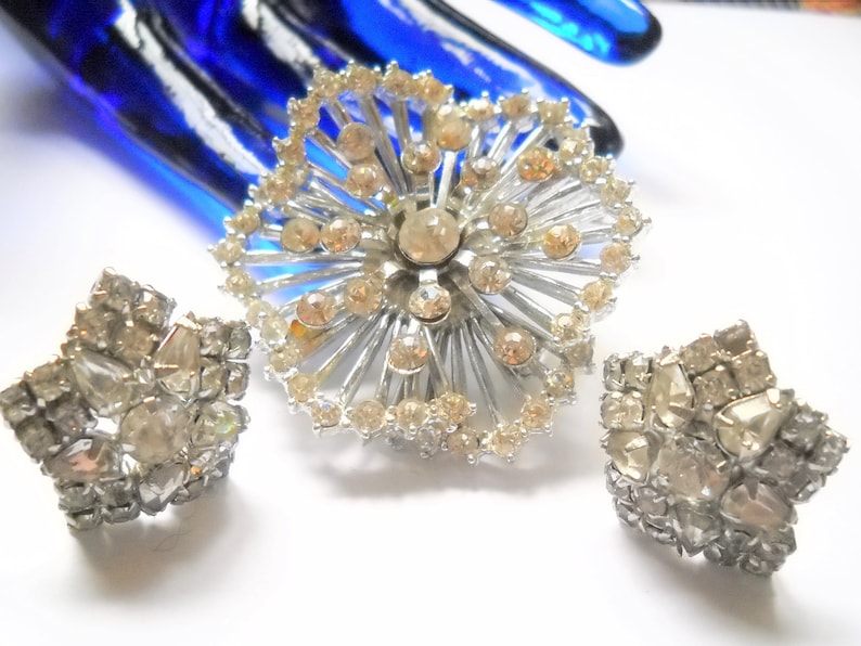 May include: A silver tone brooch and matching earrings set with clear rhinestones. The brooch is a starburst design with a central rhinestone. The earrings are star shaped.