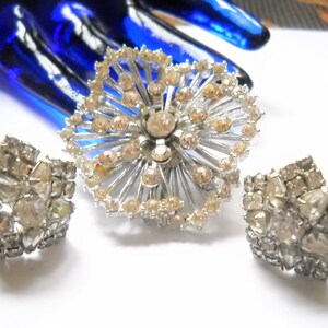 May include: A silver tone brooch and a pair of earrings with clear and light brown rhinestones. The brooch is a large, circular design with a starburst pattern. The earrings are smaller and have a cluster of rhinestones.