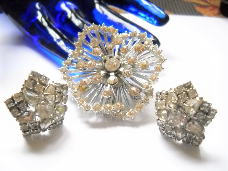 May include: A silver tone brooch and a pair of earrings with clear and light brown rhinestones. The brooch is a large, circular design with a starburst pattern. The earrings are smaller and have a cluster of rhinestones.