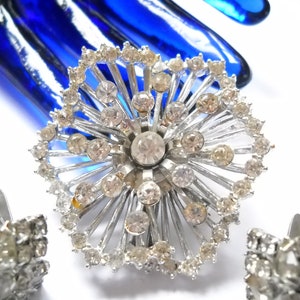May include: A silver tone brooch with a large rhinestone center and smaller rhinestones around the edge. The brooch is shaped like a flower with a starburst design.