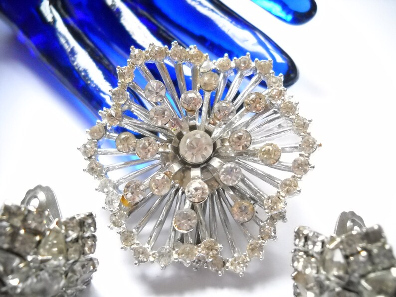 May include: A silver tone brooch with a large rhinestone center and smaller rhinestones around the edge. The brooch is shaped like a flower with a starburst design.