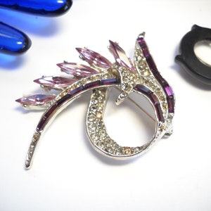 May include: A silver tone brooch with a swirl design. The brooch is decorated with clear and purple rhinestones.