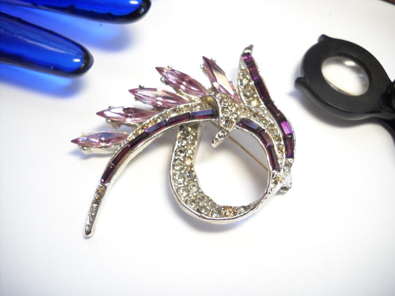 May include: A silver tone brooch with a swirl design. The brooch is decorated with clear and purple rhinestones.