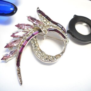 May include: A silver tone brooch with a swirl design. The brooch is decorated with clear and pink rhinestones and purple baguette-shaped stones.