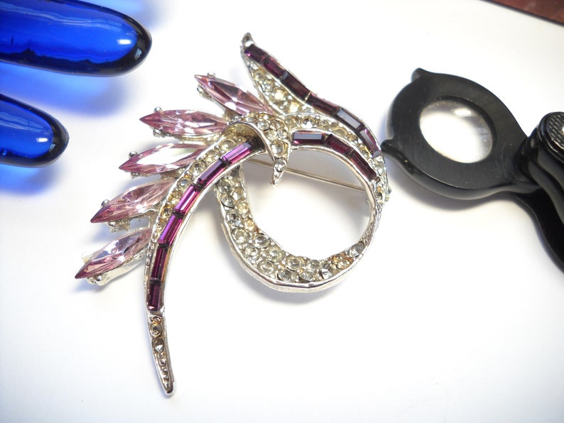 May include: A silver tone brooch with a swirl design. The brooch is decorated with clear and pink rhinestones and purple baguette-shaped stones.