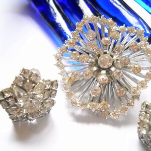 May include: A set of three silver tone brooches with clear rhinestone accents. The largest brooch is a starburst design with a central rhinestone. The smaller brooches are star shaped.