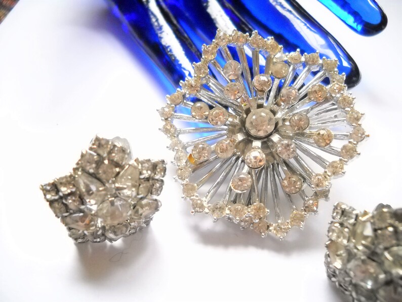 May include: A set of three silver tone brooches with clear rhinestone accents. The largest brooch is a starburst design with a central rhinestone. The smaller brooches are star shaped.