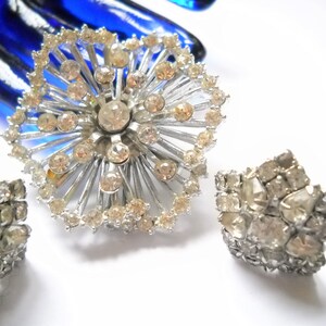 May include: A silver brooch and a pair of earrings with clear rhinestones. The brooch is shaped like a starburst with rhinestones around the edges and in the center. The earrings are also starburst shaped with rhinestones.