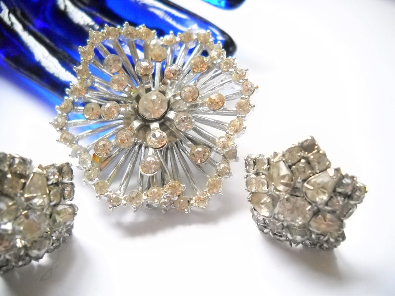 May include: A silver brooch and a pair of earrings with clear rhinestones. The brooch is shaped like a starburst with rhinestones around the edges and in the center. The earrings are also starburst shaped with rhinestones.