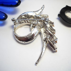 May include: Silver tone brooch featuring a dragon design. The dragon is decorated with clear and amber coloured rhinestones. The brooch has a pin back.
