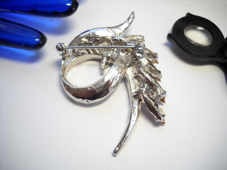 May include: Silver tone brooch featuring a dragon design. The dragon is decorated with clear and amber coloured rhinestones. The brooch has a pin back.