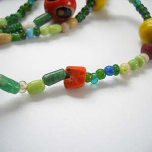 Op de afbeelding: Een kleurrijke kralenketting met groene, rode, gele en witte kralen. De ketting heeft verschillende vormen en maten kralen, waaronder ronde, vierkante en rechthoekige kralen.